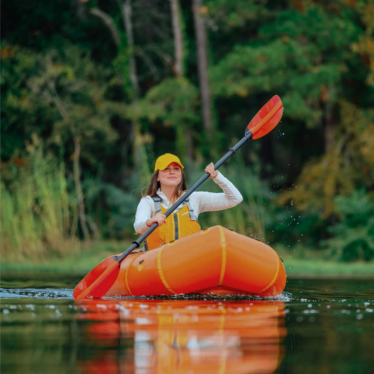 A smiling woman paddling an orange Advanced Elements inflatable packraft on a calm lake surrounded by lush green trees.