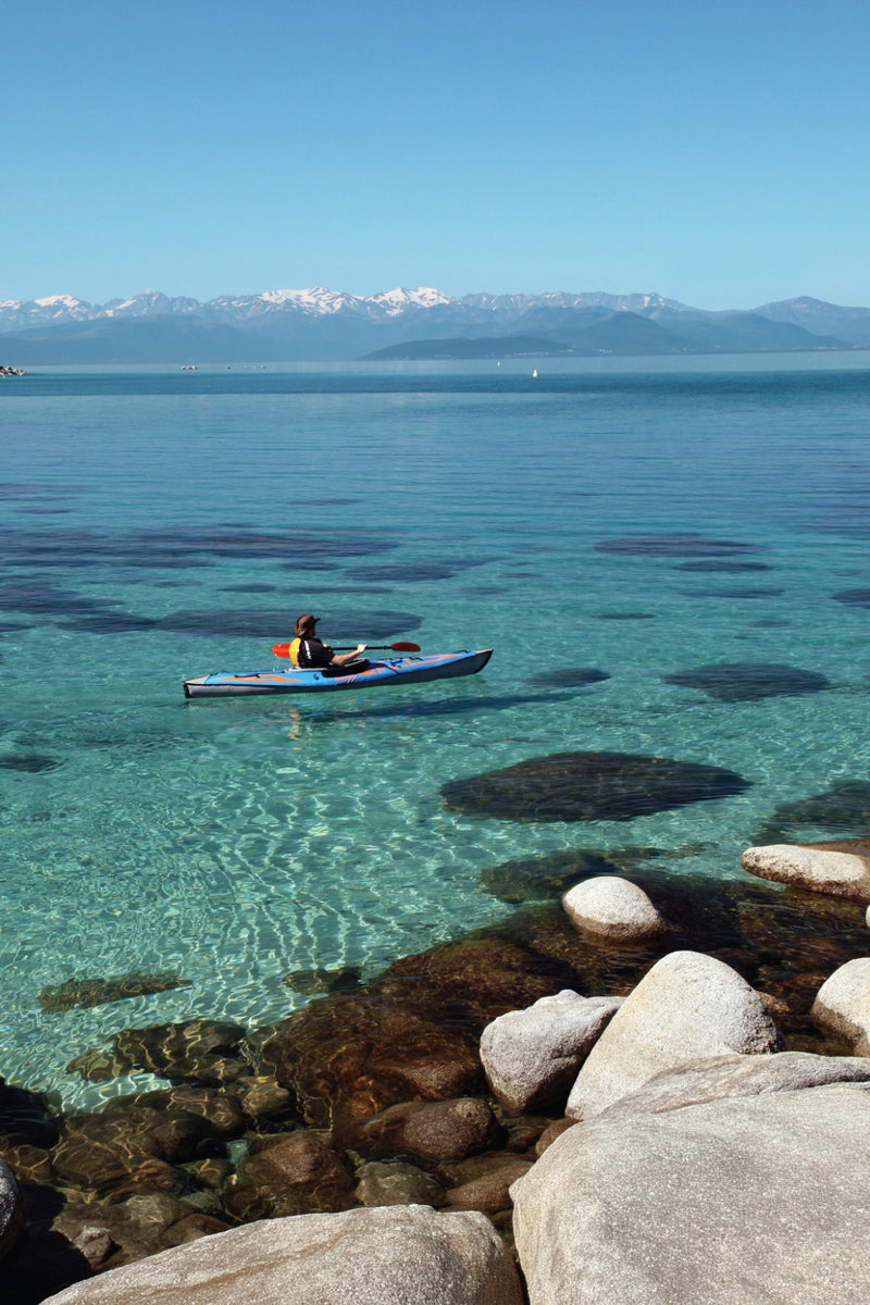 A person paddling an Advanced Elements inflatable kayak from the AdvancedFrame Series on crystal-clear turquoise water with snow-capped mountains in the background.