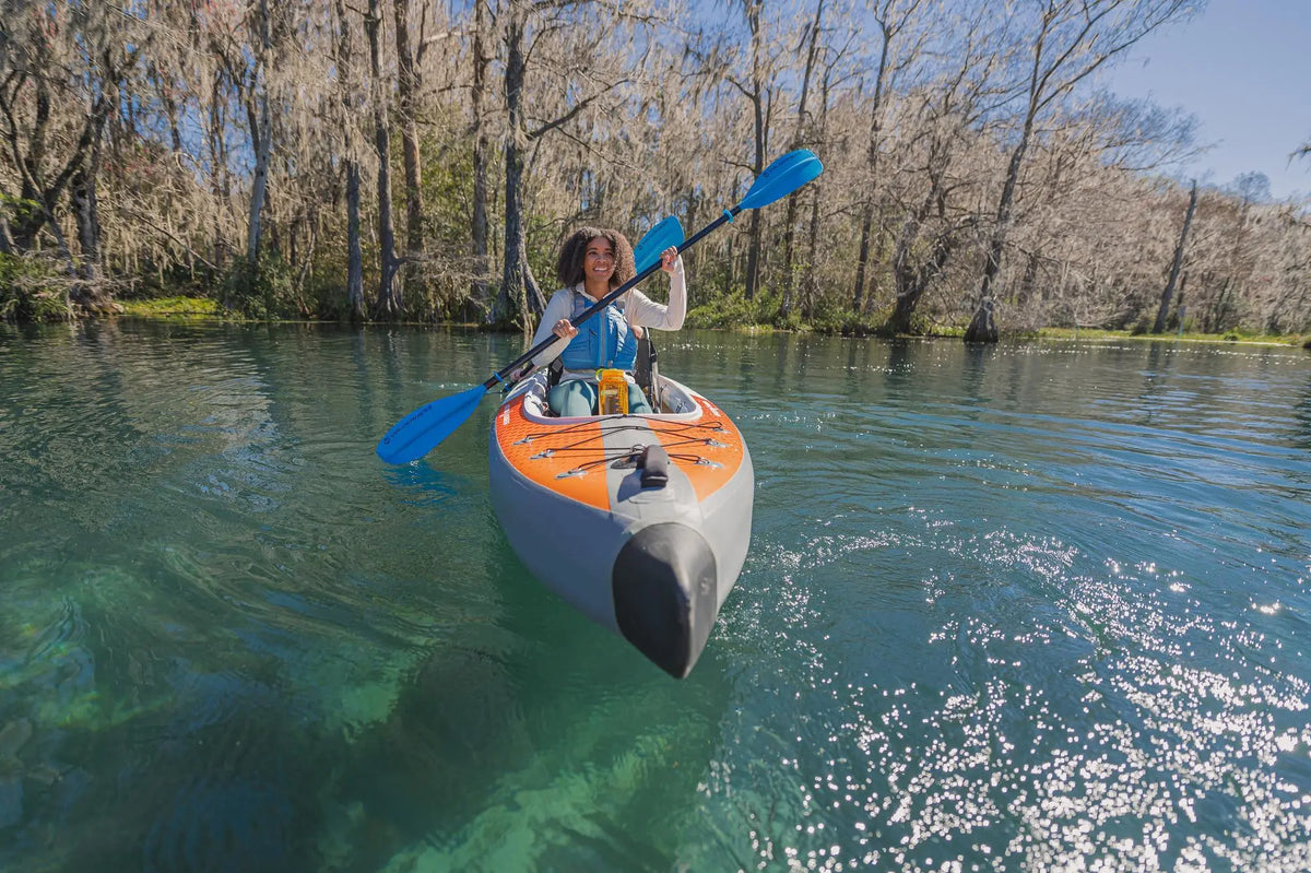 A person paddling an orange and grey Advanced Elements Airvolution inflatable kayak on clear water surrounded by trees.