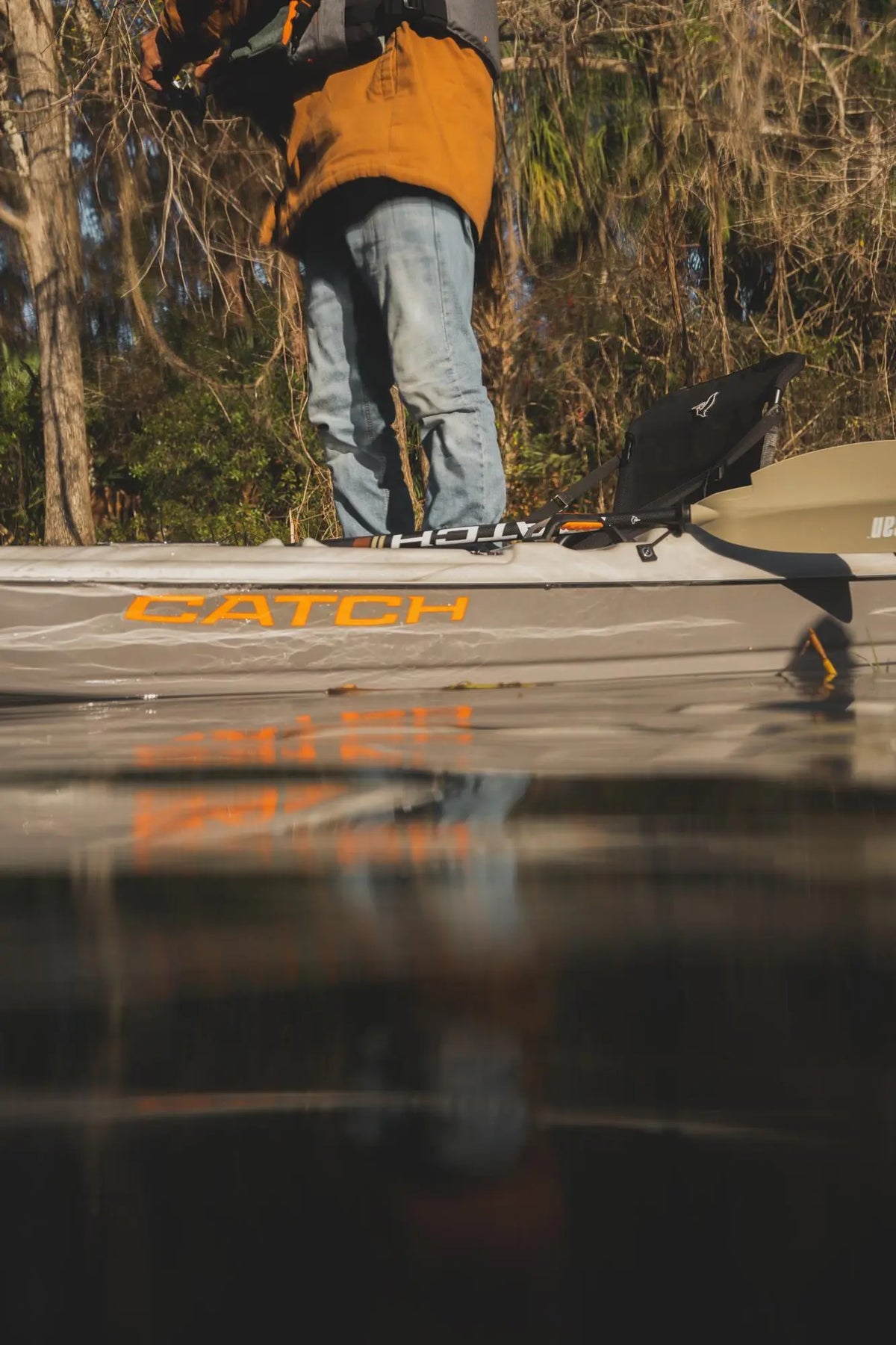 Low-angle shot of an angler standing on a Pelican CATCH fishing kayak on water with reflections.