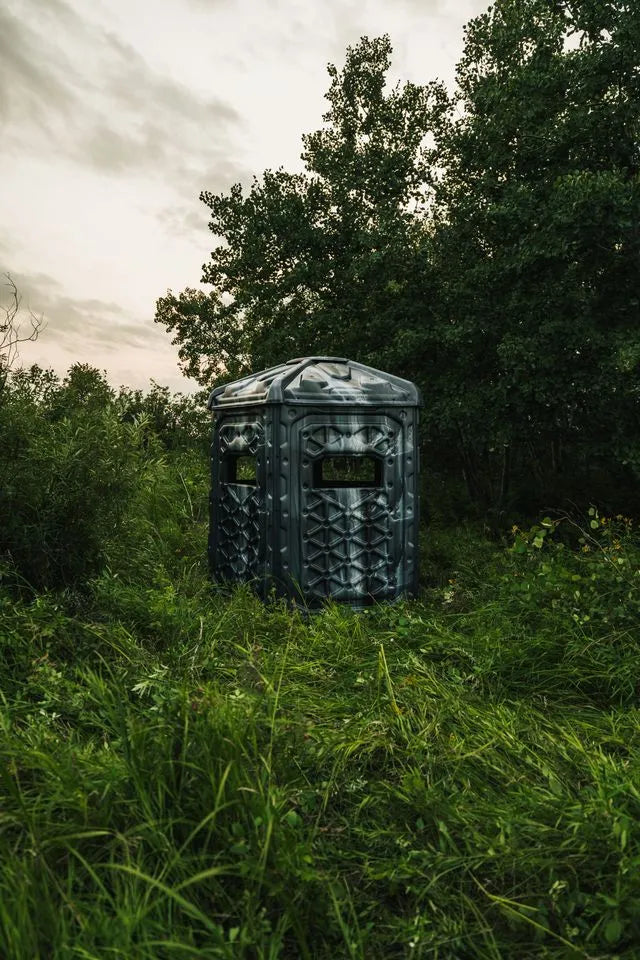 A hard-shell camouflage Pelican CATCH hunting blind positioned in tall green grass surrounded by dense trees under an overcast sky.