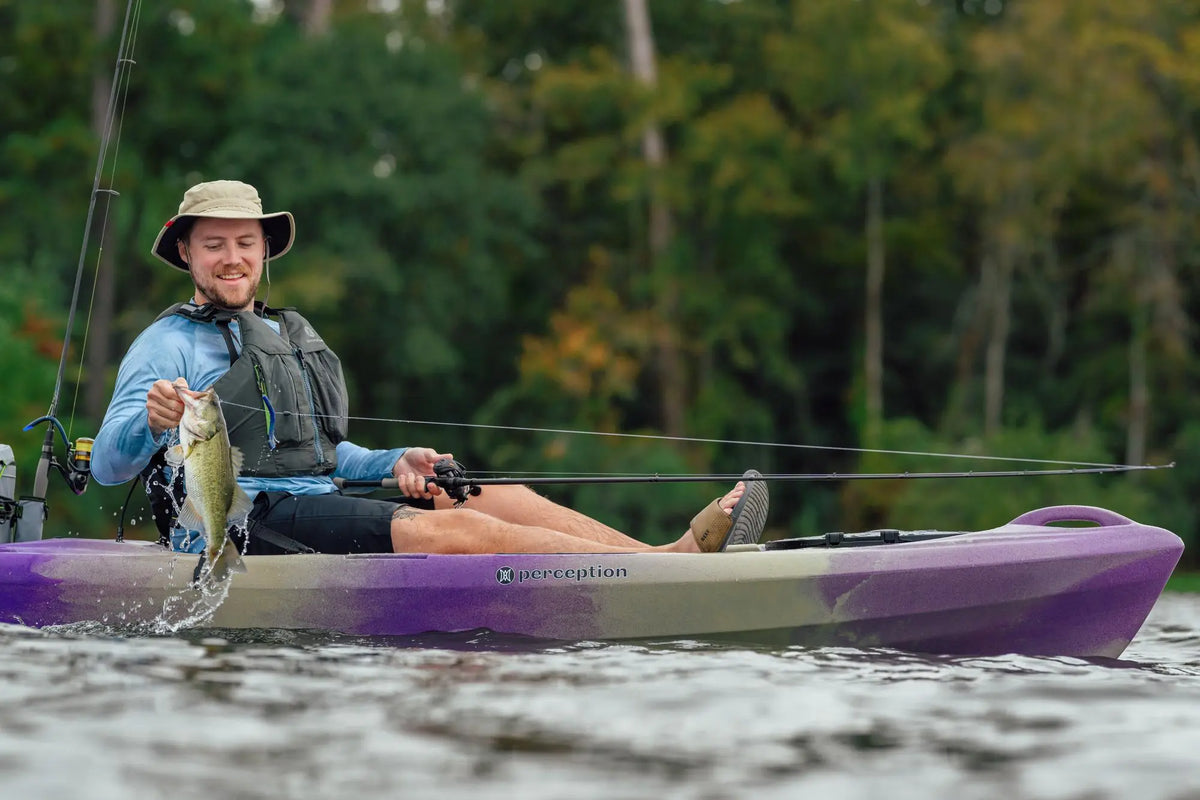 A smiling angler holding up a caught fish on a purple Perception Pescador kayak.