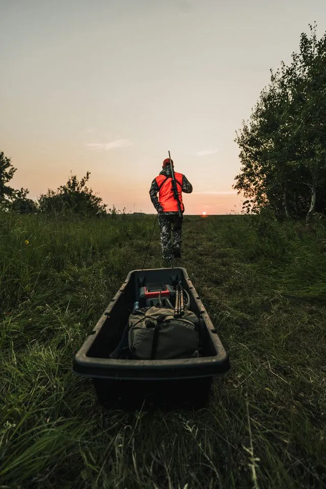 A hunter in high-visibility safety gear walking through a field at sunrise next to a utility sled.